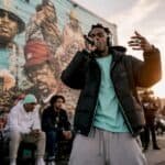 Stevie The Manager rapping outdoors with a crowd and mural in background.