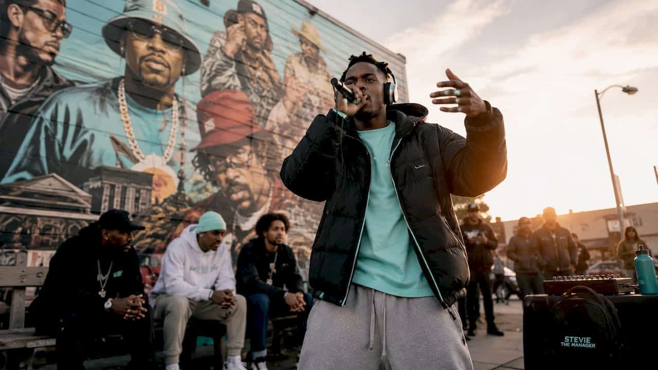 Stevie The Manager rapping outdoors with a crowd and mural in background.